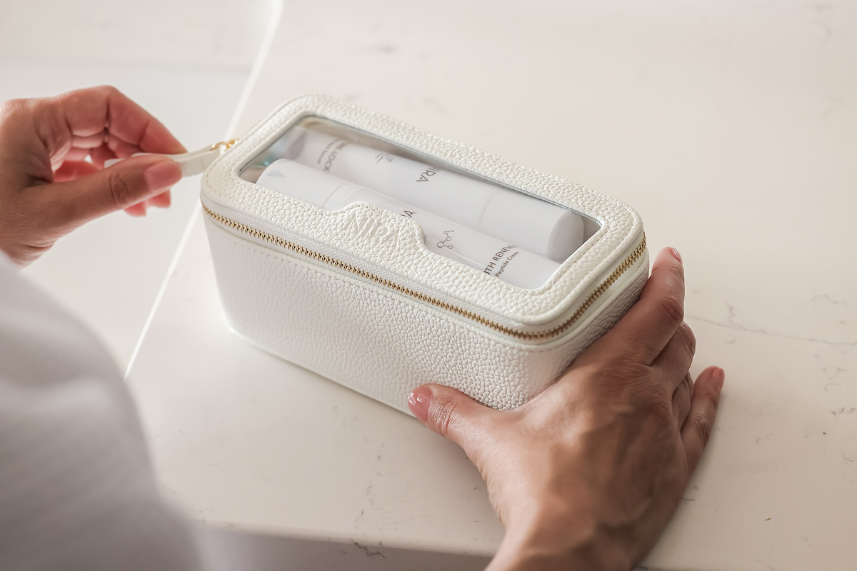 Close-up of a person's hands placing or removing two white skincare bottles from a sleek, white zippered travel or cosmetics bag with a clear top window, sitting on a light-colored countertop.