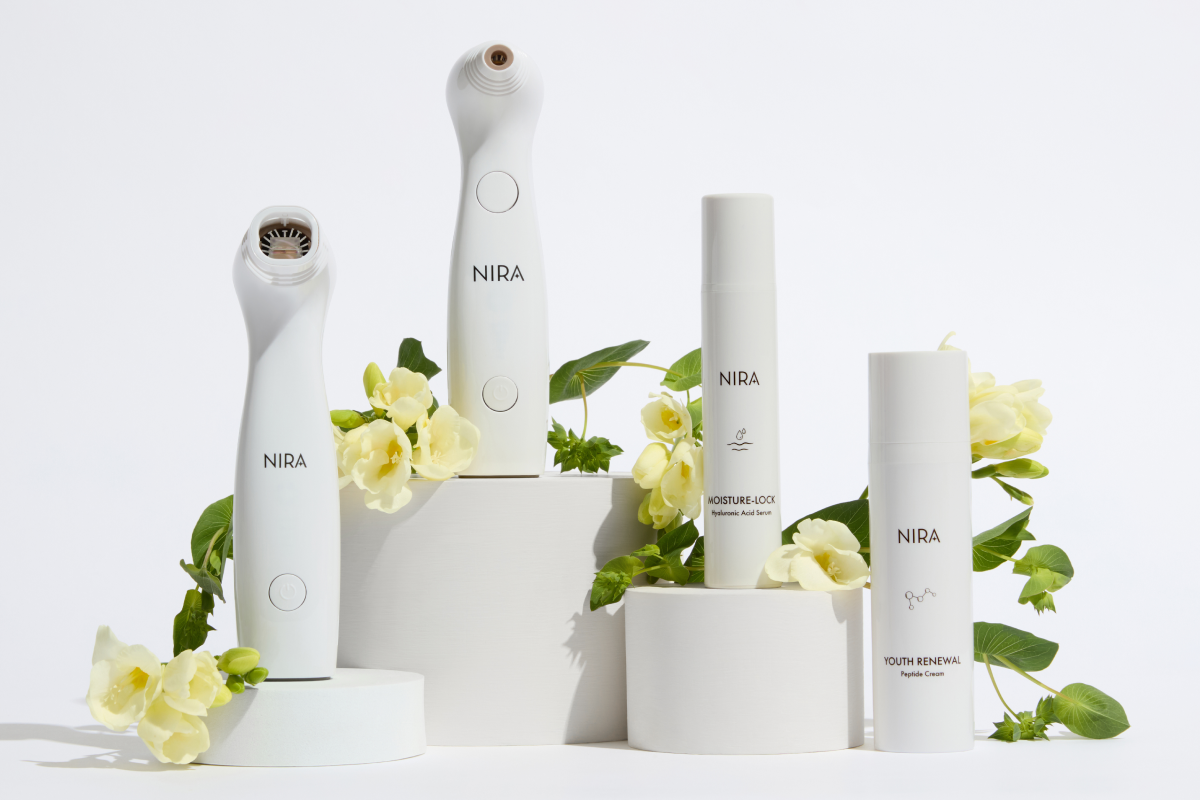 Studio shot featuring four white NIRA skincare products arranged on a tiered display of white geometric blocks, surrounded by pale yellow flowers and green leaves. The products include two handheld laser devices and two bottles of NIRA skincare products.