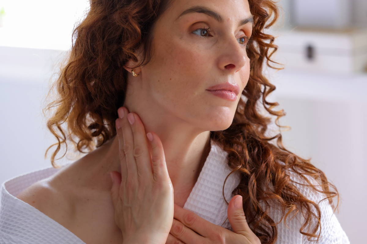 A woman with curly red hair, wearing a white robe, gently touches her neck and chest. She looks off to the side with a serene expression.