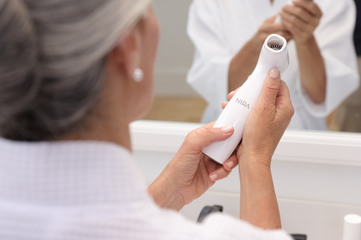 Over-the-shoulder view of a woman with gray hair and a white bathrobe, holding a white NIRA skincare laser device and examining it in the reflection of a bathroom mirror. The focus is on the device and the woman's hands.