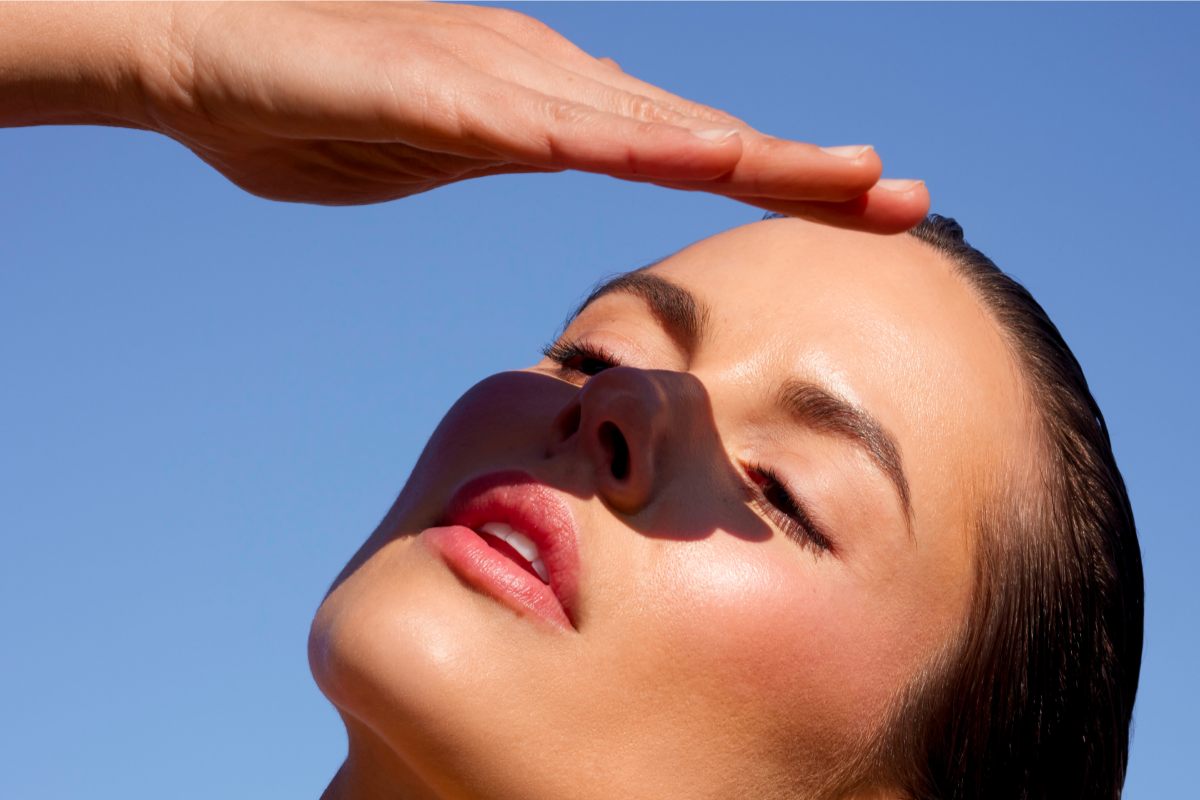 A sunlit close-up of a woman's face and neck against a clear blue sky. She has glowing, dewy skin and is shading her forehead with her hand.