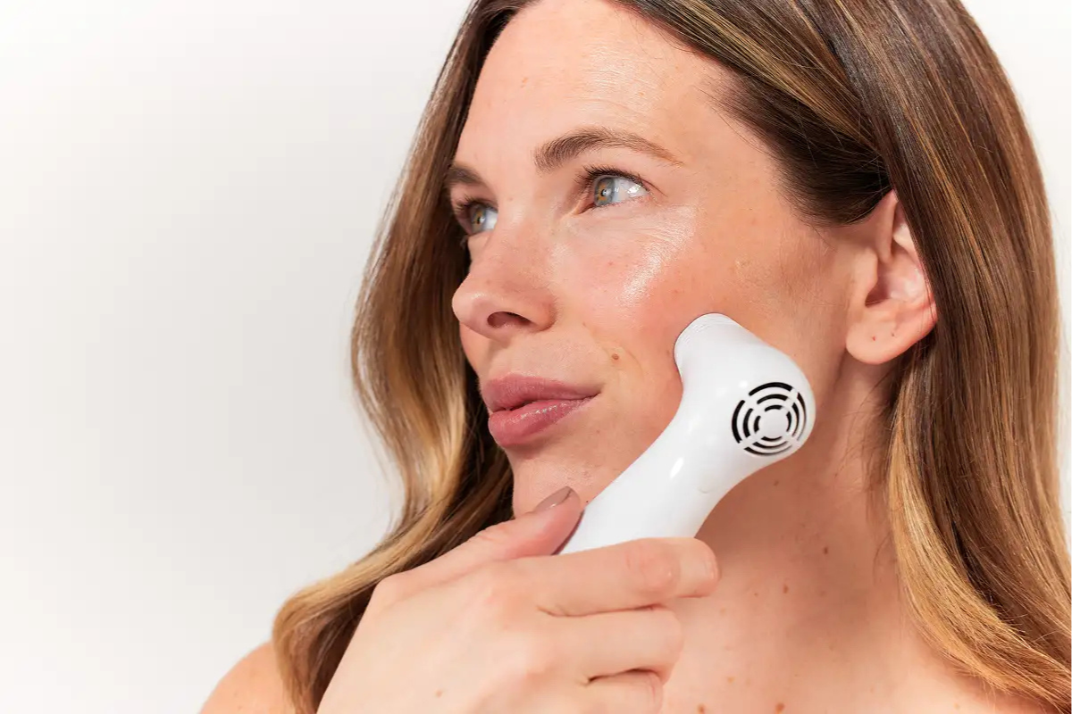 A close-up shot of a woman using a white, handheld NIRA Skin Laser device on her cheek near her temple.