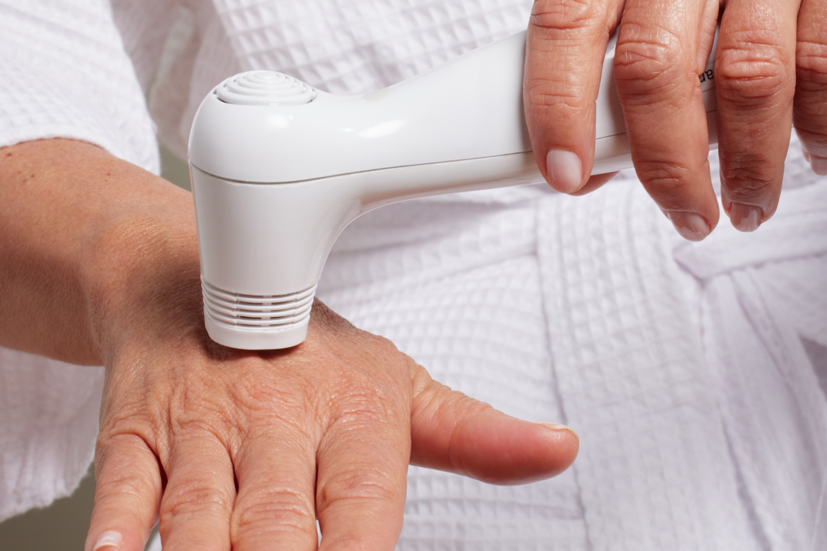 A close-up of a person wearing a white robe, holding a white Nira laser and using it on the back of their hand.