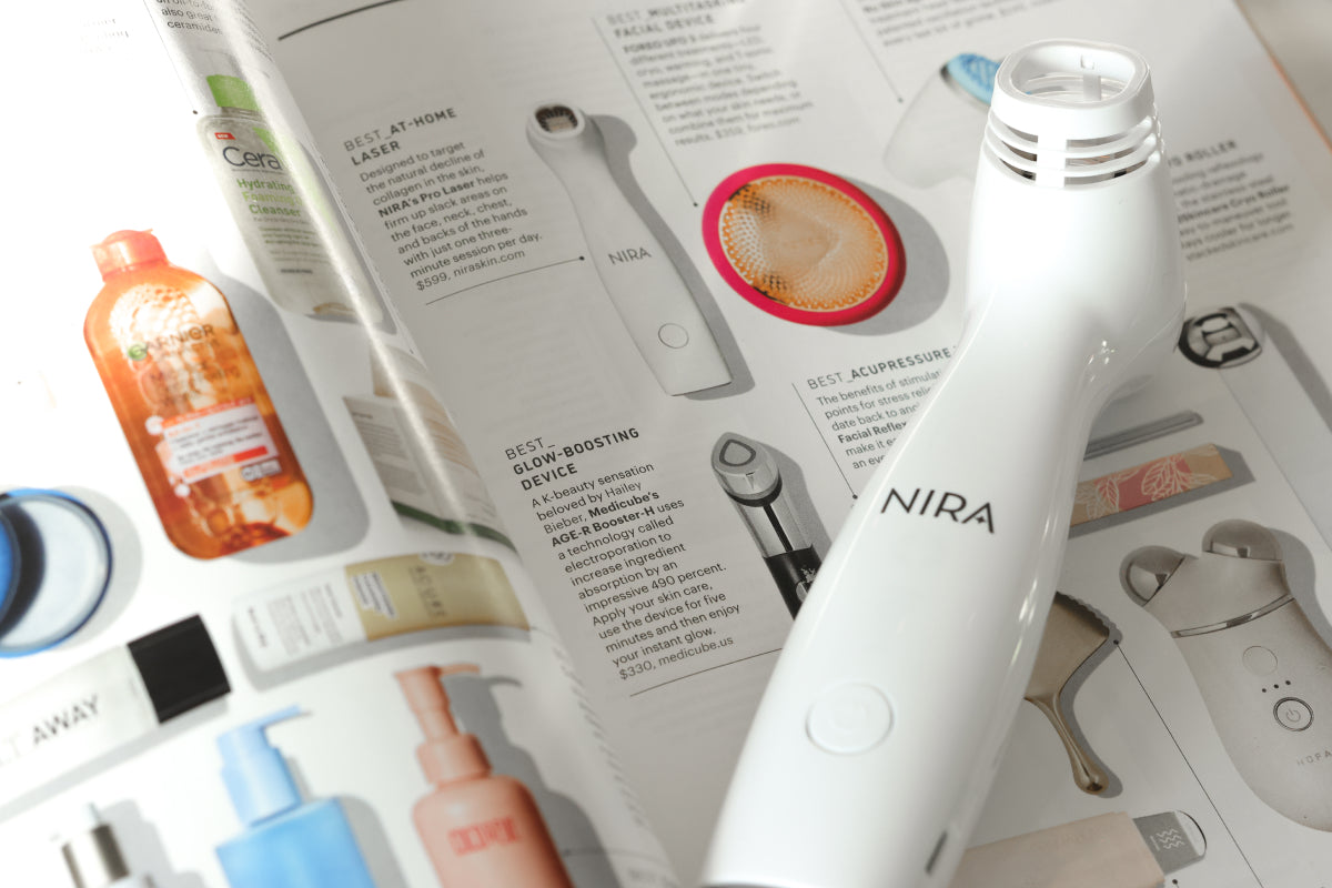 A white Nira skincare laser is placed on an open magazine spread featuring various skincare devices and products. Other blurred skincare items are visible in the background.