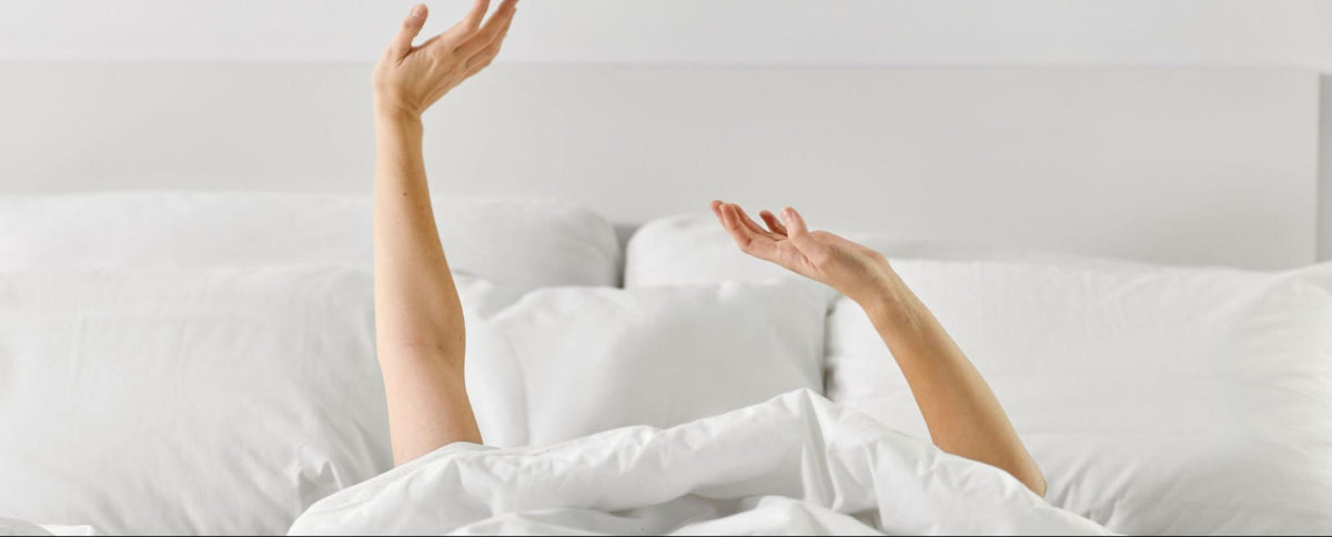 person with face covered hands in the air on a bed surrounded by white covers and pillows 