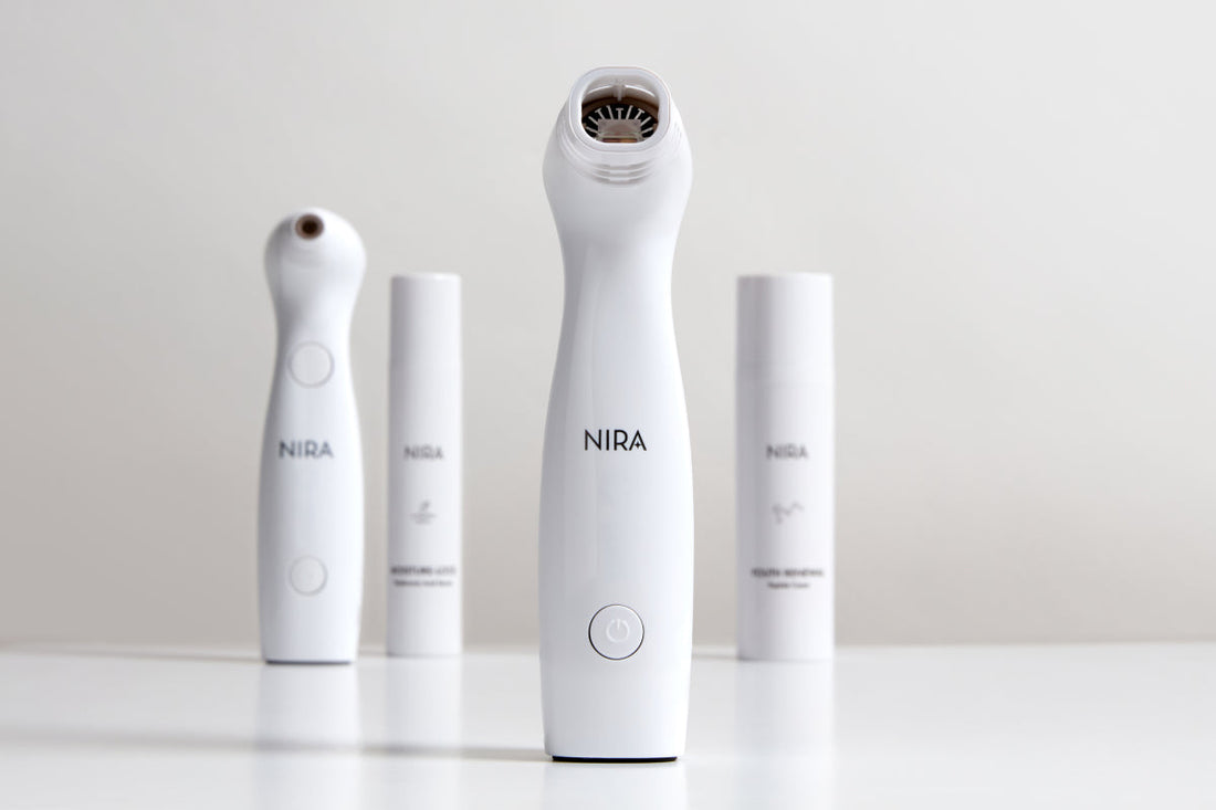 A brightly lit, professional studio shot features four white NIRA skincare devices and products on a reflective surface. The NIRA Pro Laser device is in the center foreground, showing a handheld unit with a black treatment head.