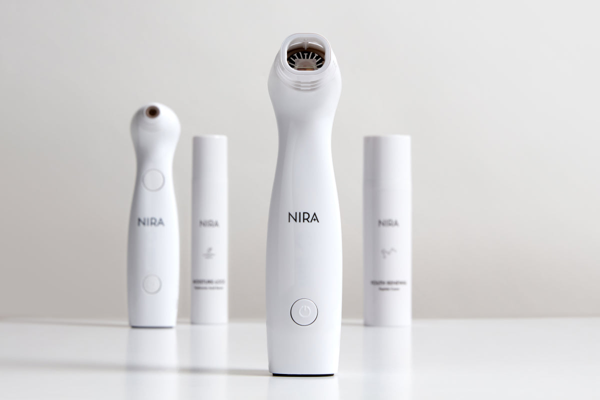 A brightly lit, professional studio shot features four white NIRA skincare devices and products on a reflective surface. The NIRA Pro Laser device is in the center foreground, showing a handheld unit with a black treatment head.