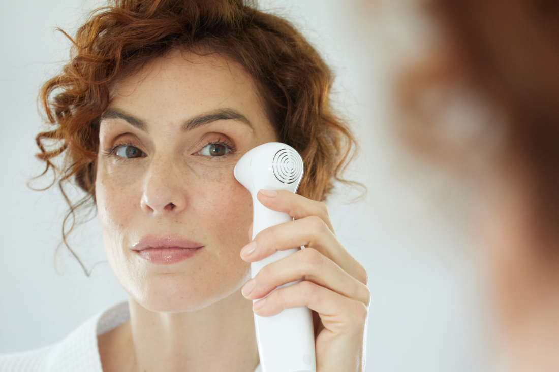 A red-haired woman with fair skin, wearing a white robe, holds a NIRA at-home laser device to her right cheek.