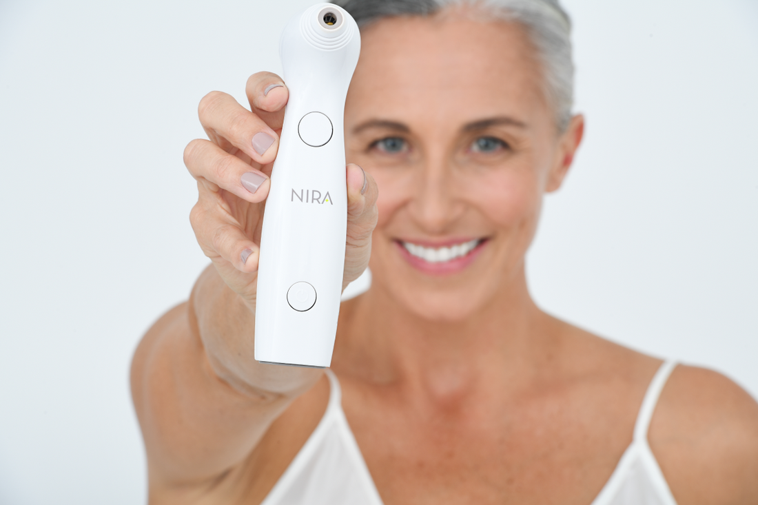 A middle-aged woman with gray hair and a white tank top is holding a white NIRA skincare laser device directly toward the camera. The device is in sharp focus in the foreground, while the smiling woman's face is slightly blurred in the background.