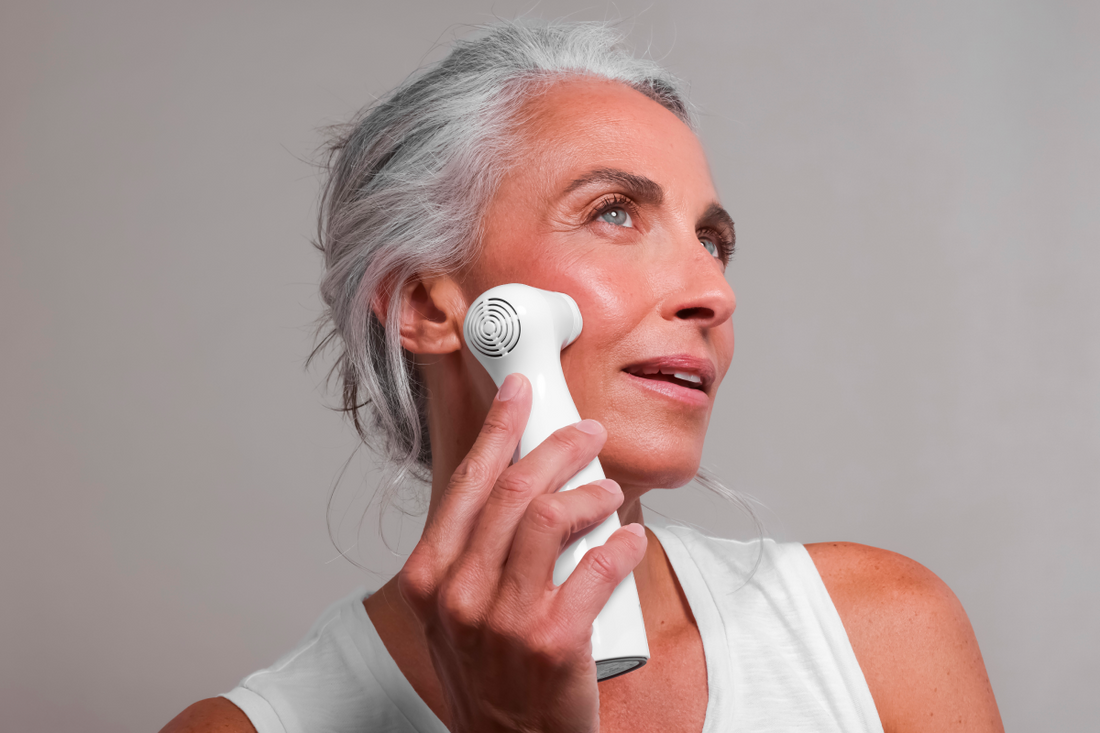 Close-up of an older woman with striking gray hair and a white tank top, looking up and to the side as she applies a white handheld skincare laser device to her cheek. She has a subtle blush on her cheek, set against a plain light gray background.