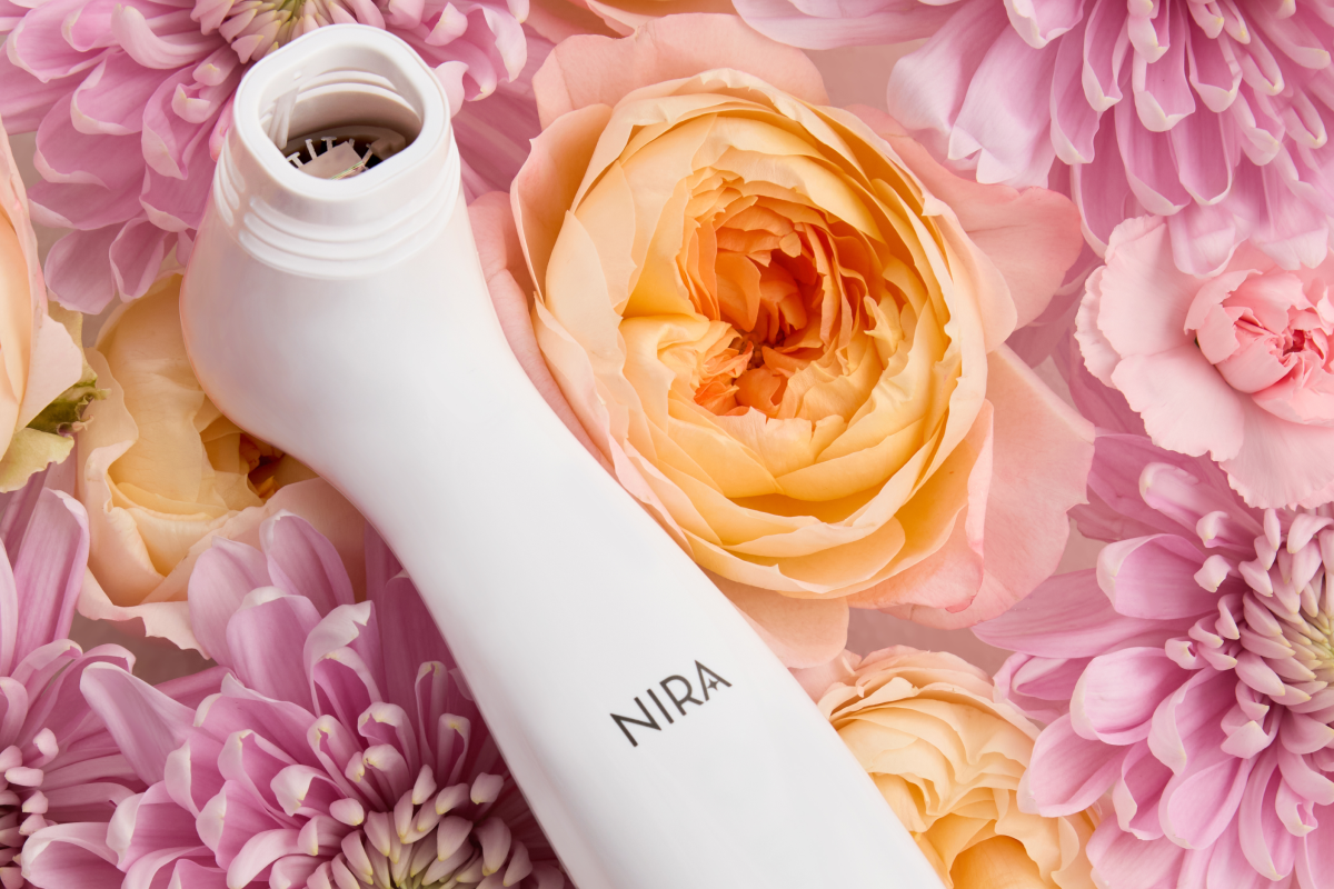 Close-up, overhead shot of a white handheld skincare laser device, branded with "NIRA," resting on a bed of pink and peach flowers, including roses and chrysanthemums. The applicator tip of the device is visible at the top.