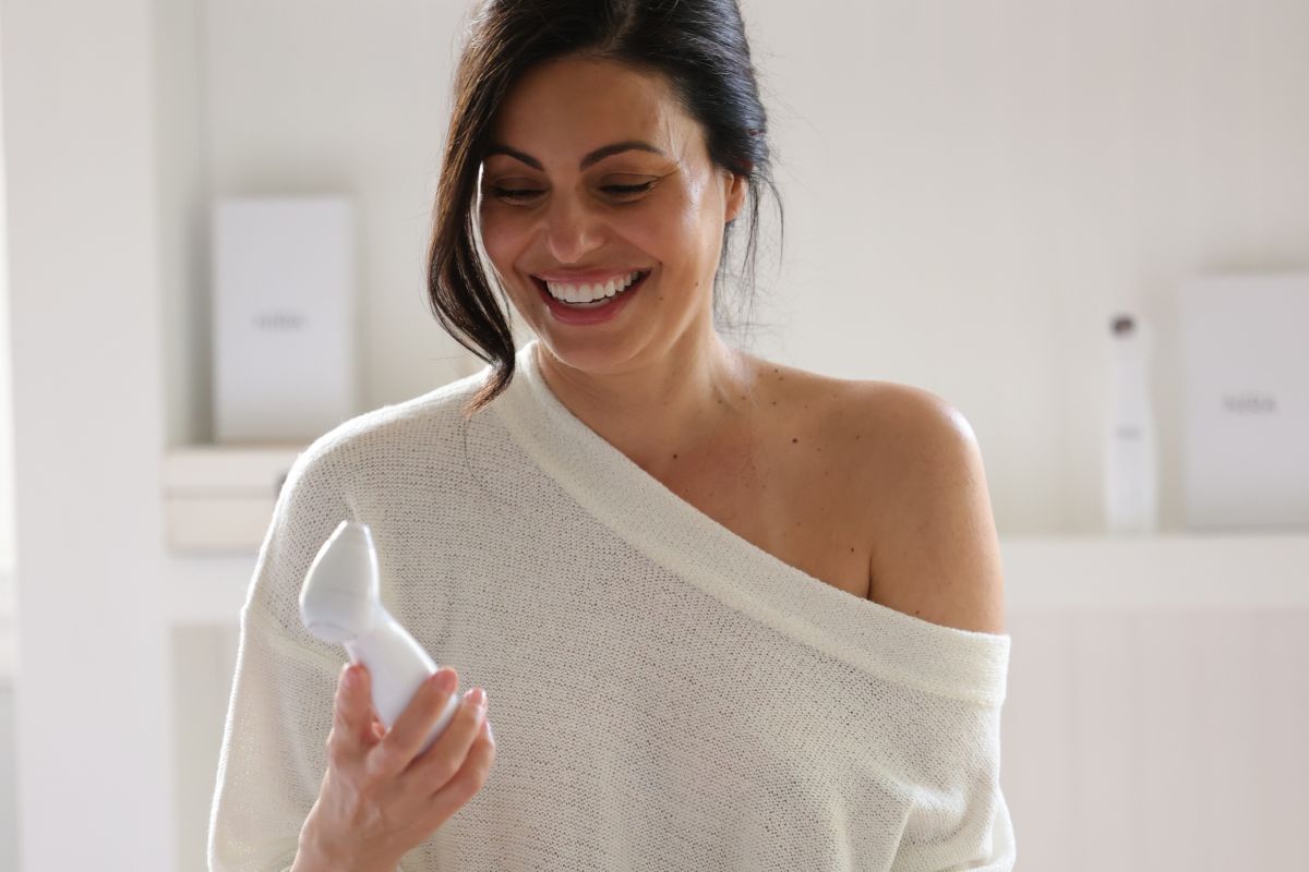 A smiling woman with dark hair in a casual, off-the-shoulder white top is sitting on a bed, holding and looking down at a white, handheld NIRA Skin Laser device. Other NIRA devices are visible on the bed in the foreground.
