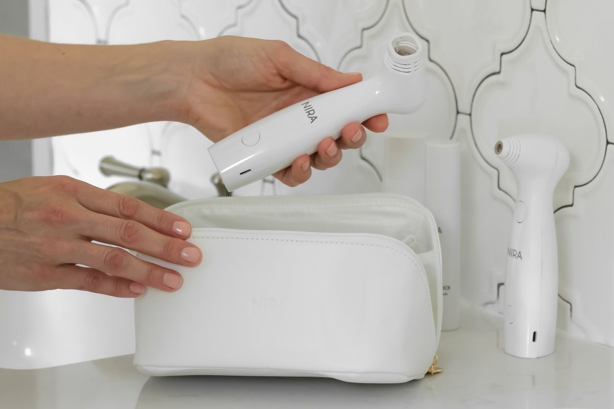 A close-up of a person's hands holding a NIRA Pro Laser over a white makeup bag with a clear top, next to a second similar device, all resting on a white bathroom vanity with a white tiled backsplash.