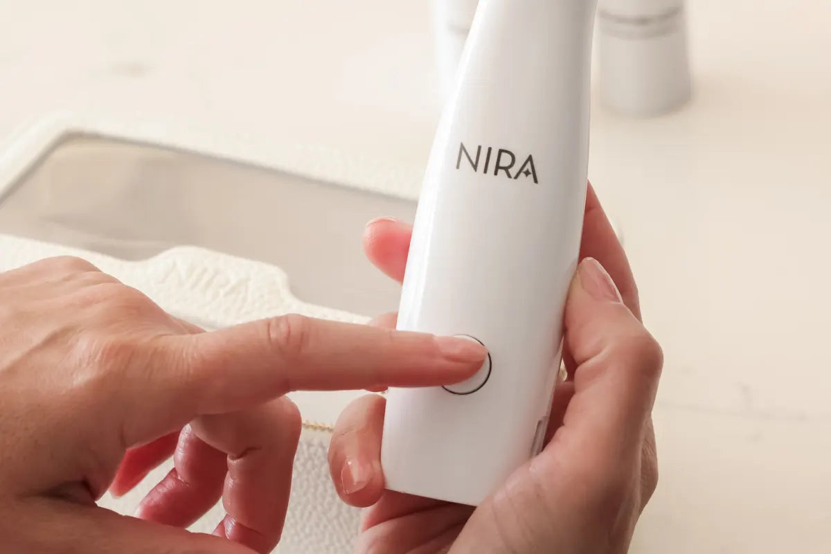 Close-up of a person's hands holding a white NIRA skin-care laser device, with a finger pressing the power button.