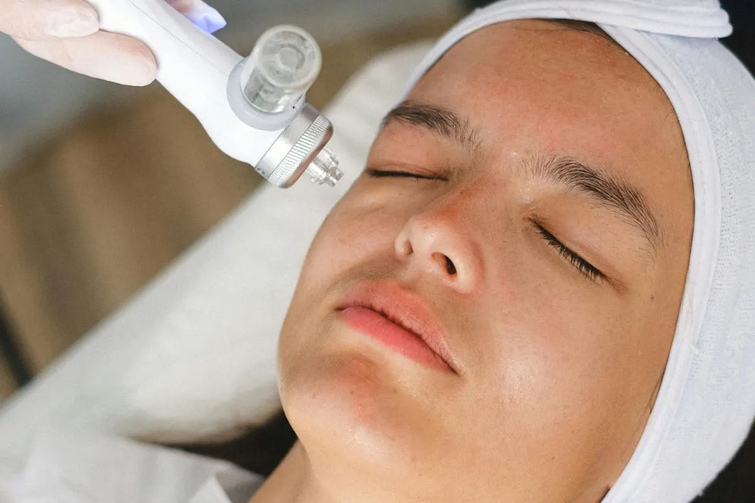 A young woman with her eyes closed is receiving an in-house laser or light-based skin treatment on her cheek, performed by a technician in white gloves using a handheld white device.