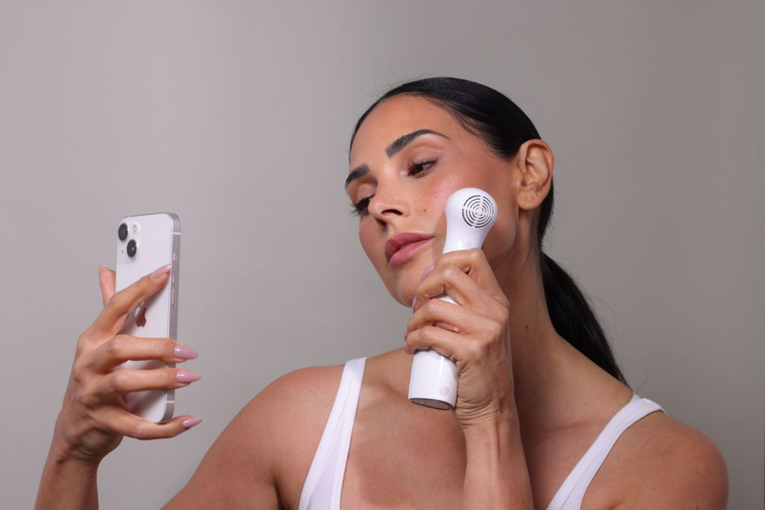 Model using NIRA Pro at-home laser device, holding phone camera for selfie, demonstrating skin rejuvenation and at-home skincare routine.