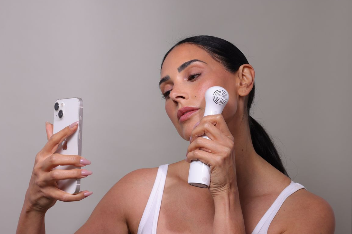 Model using NIRA Pro at-home laser device, holding phone camera for selfie, demonstrating skin rejuvenation and at-home skincare routine.