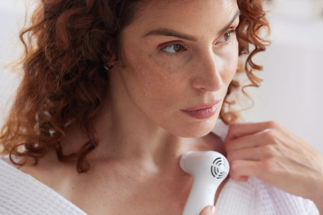 Model in robe looking intently into mirror while using NIRA Pro at-home laser device, demonstrating skincare routine and skin rejuvenation results.