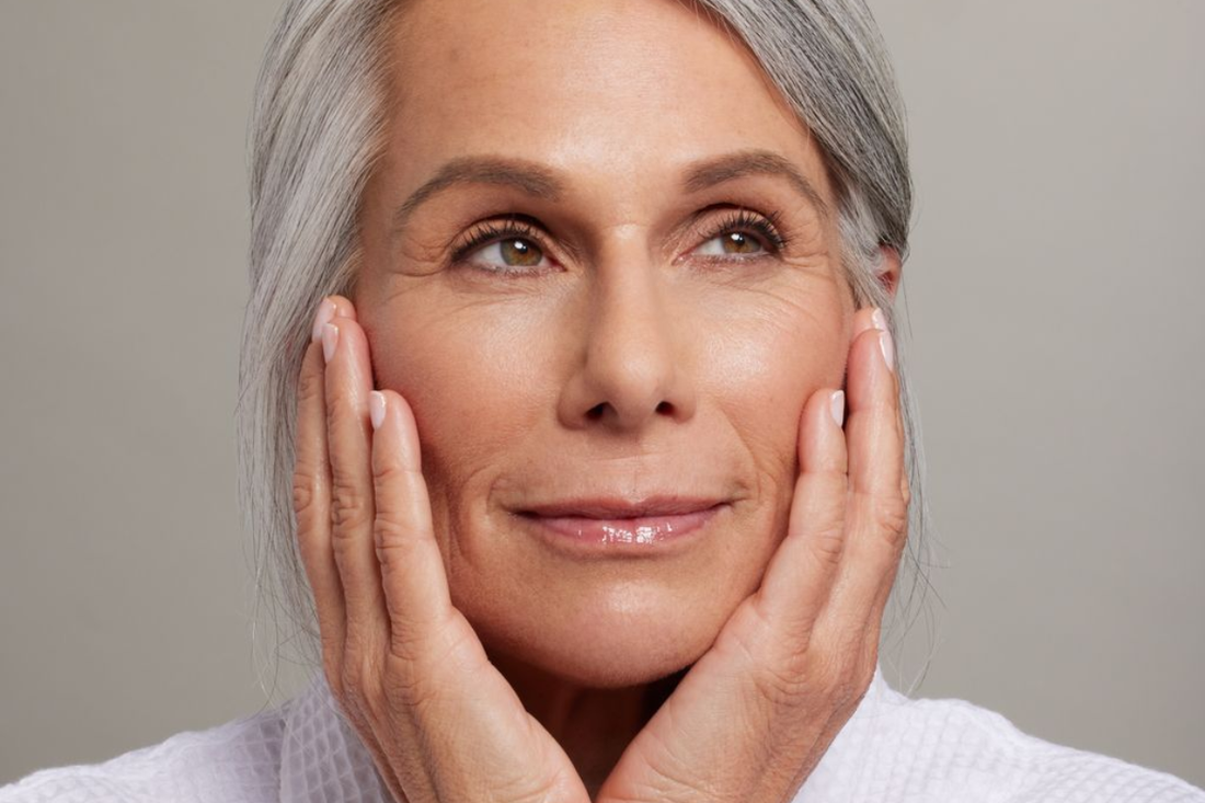 Elegant older woman with gray hair resting her face in her hands, highlighting smooth, radiant mature skin.