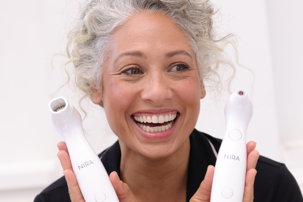 Smiling woman with gray hair holding the NIRA Pro and Precision Lasers, showcasing at-home devices for collagen renewal and youthful skin.