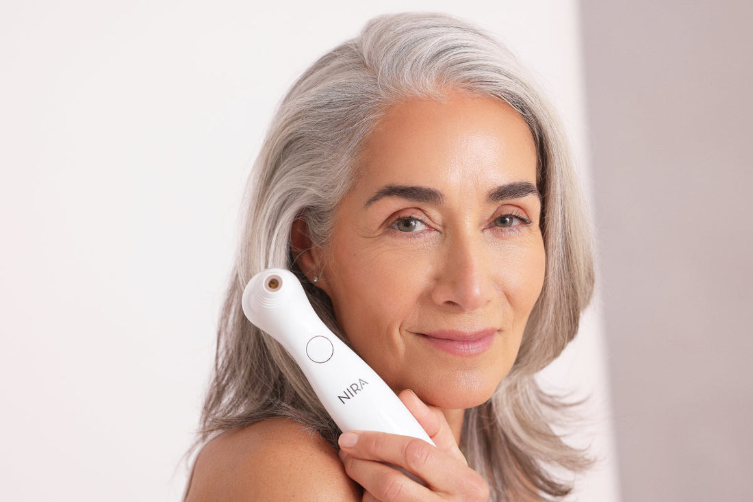 A mature woman with silver hair is holding a white, handheld NIRA laser device up to the side of her face near her temple. She is looking directly at the camera with a calm expression. The background is a soft, light gray.
