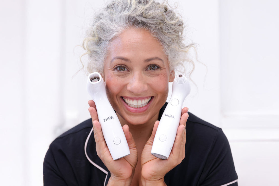 A smiling woman with curly gray hair, wearing a black top, holds up two white Nira skincare lasers on either side of her face.