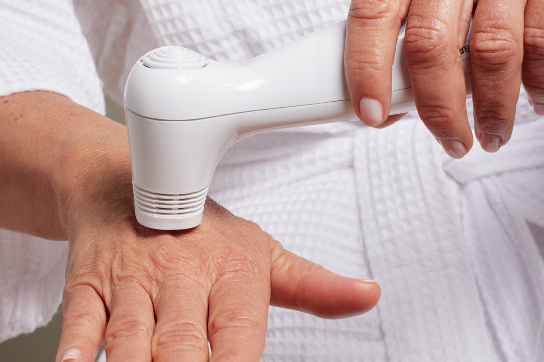 A close-up of a person wearing a white robe, holding a white Nira laser and using it on the back of their hand.