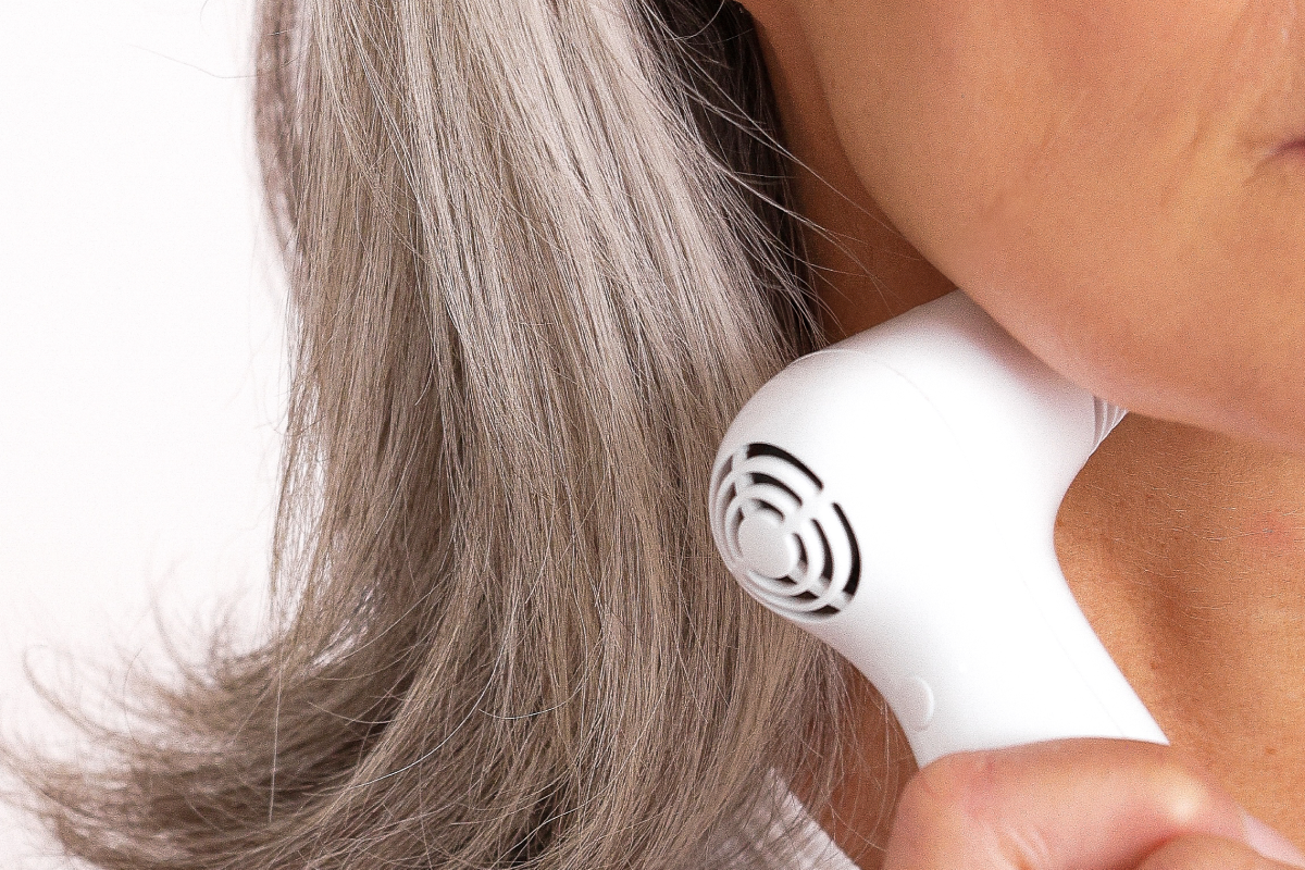 A close-up of a woman with gray hair wearing a white tank top, holding a white Nira skin laser to her neck