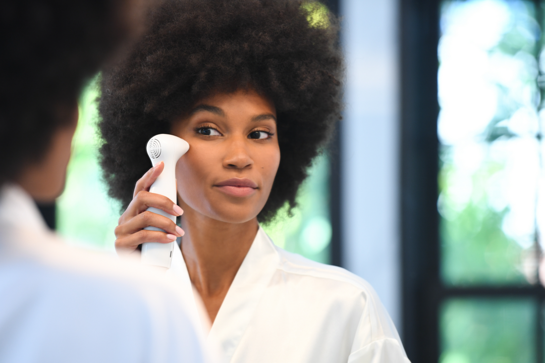 A woman wearing a white robe, looks at her reflection in a mirror while holding a white Nira laser to her temple