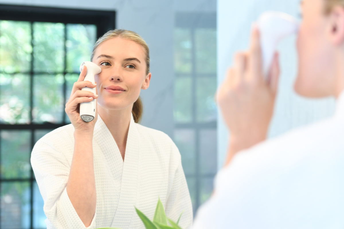 A young woman with blonde hair, pulled back into a ponytail, wears a white bathrobe and uses a NIRA skincare laser on her cheek while looking at her reflection in a mirror. A black-framed window and a green plant are visible in the background.