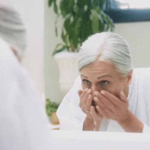 Cargar vídeo: Woman in a robe leaning toward a bathroom mirror, cleansing her face before a NIRA Pro 3 laser treatment